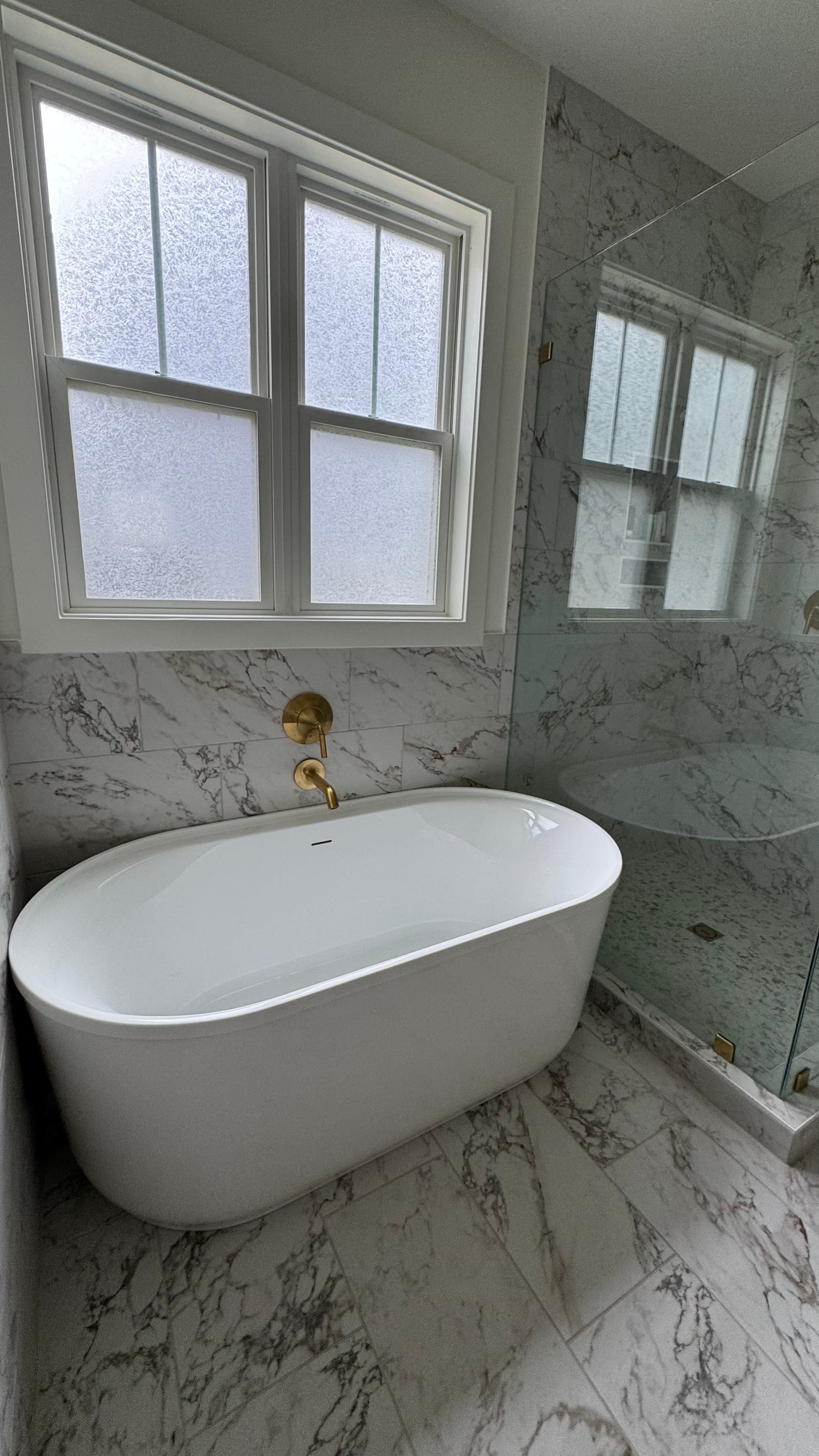 Freestanding oval tub against large-format marble-look tile, with a brushed gold wall-mounted filler and obscured-glass window